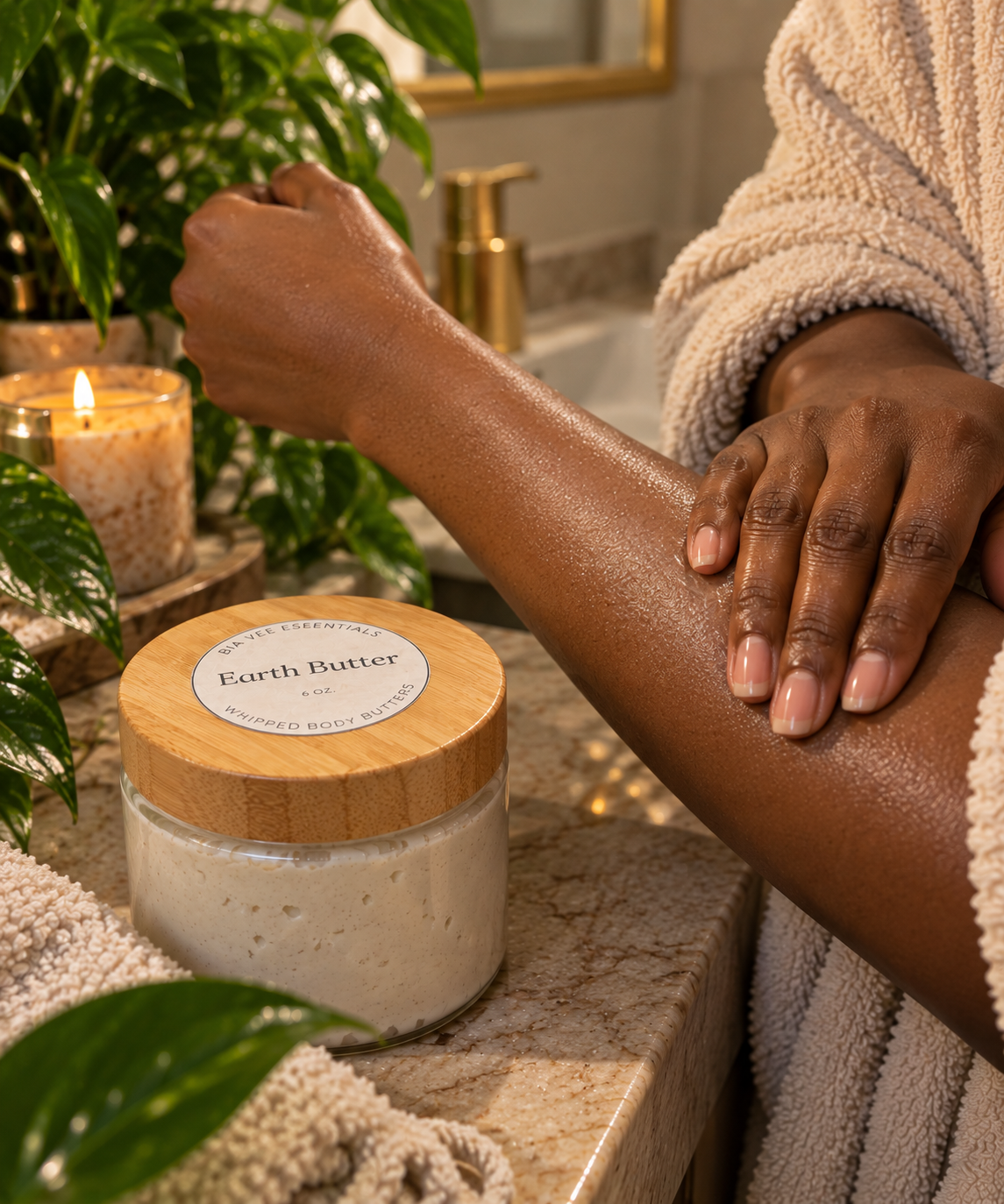 A person in a beige robe applies a cream body butter from a jar with a wooden lid labeled 'Earth Butter'. The jar sits on a marble bathroom counter next to a lit candle and green plants.